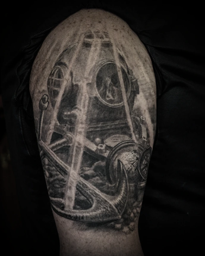 Realistic nautical tattoo on the upper arm showing a rusted diver helmet, a human skull, and a heavy anchor in black and grey shading.