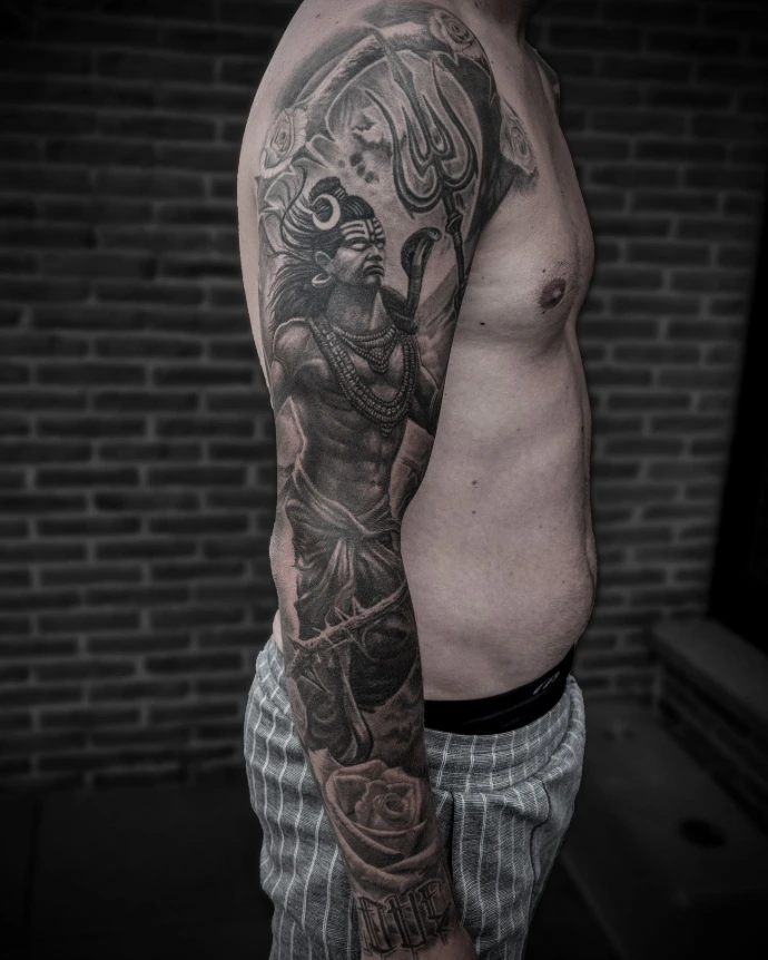 Realistic forearm tattoo of Lord Shiva (Mahadev) with intricate jewelry and smoke background, shading from shoulder to wrist.