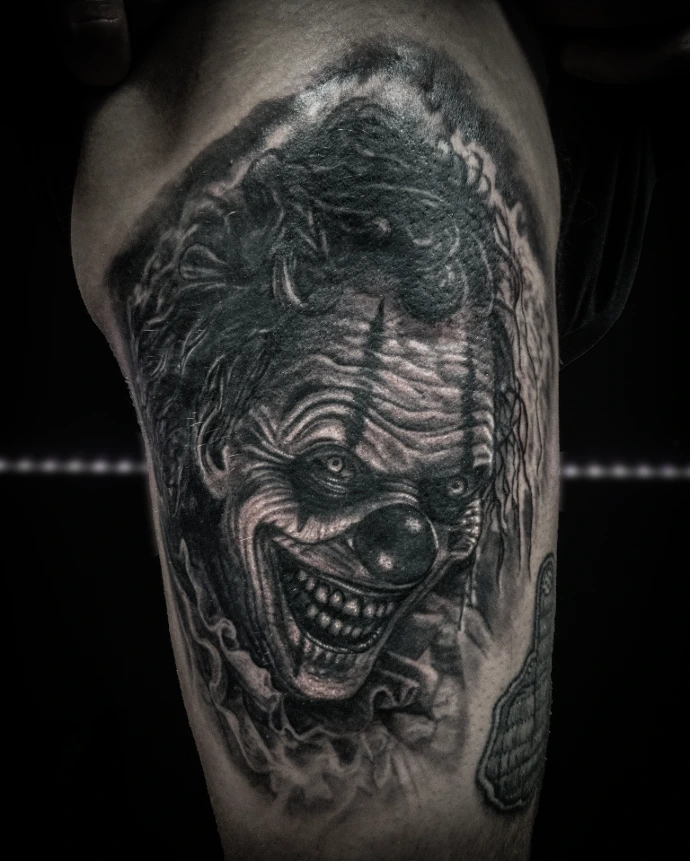 Realistic horror portrait tattoo on thigh showing a close-up of an evil clown face with high-contrast shading.