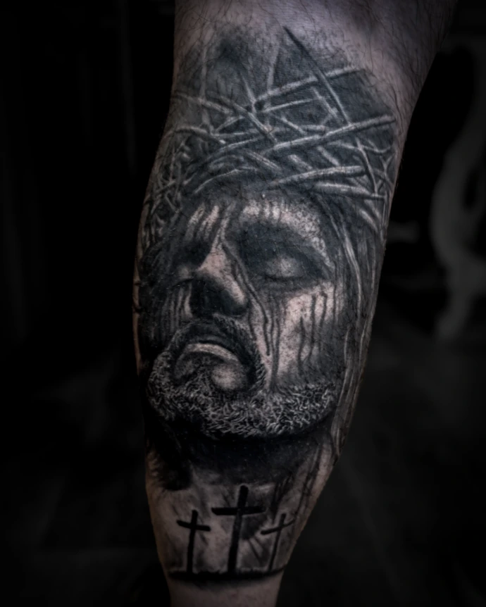 Black and grey realism tattoo of Jesus Christ wearing a crown of thorns with blood details and three crosses silhouette at the bottom.