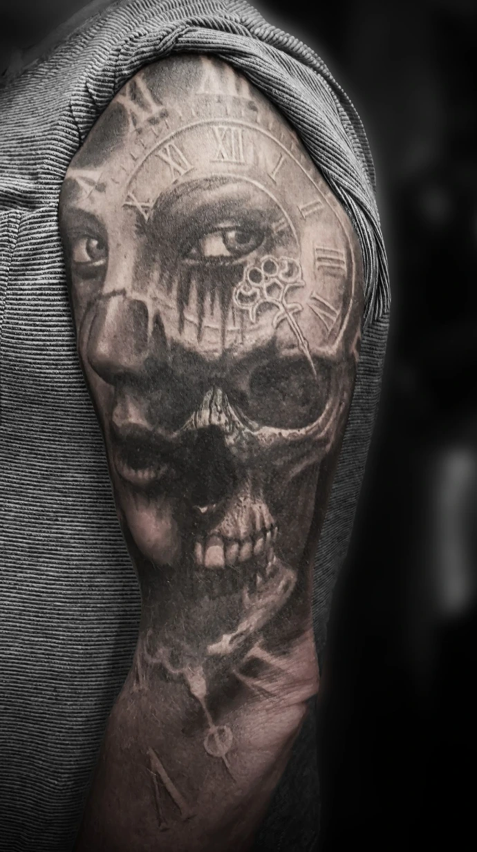 Black and grey realism tattoo on the upper arm featuring a human skull merging with a vintage clock face and a skeleton key.