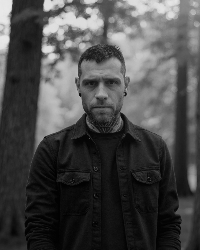 Gennaro De Schrijver Gennaro outside in a forest, looking serious and confident at the camera. Wearing a black shirt and black tshirt.